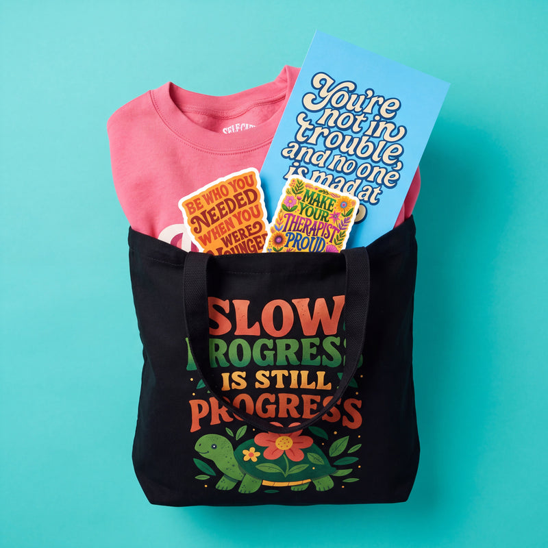 A black Slow Progress Is Still Progress tote bag filled with a self-care gift basket: a pink affirmation sweatshirt, two mental health stickers, and a You're Not in Trouble and No One Is Mad at You postcard
